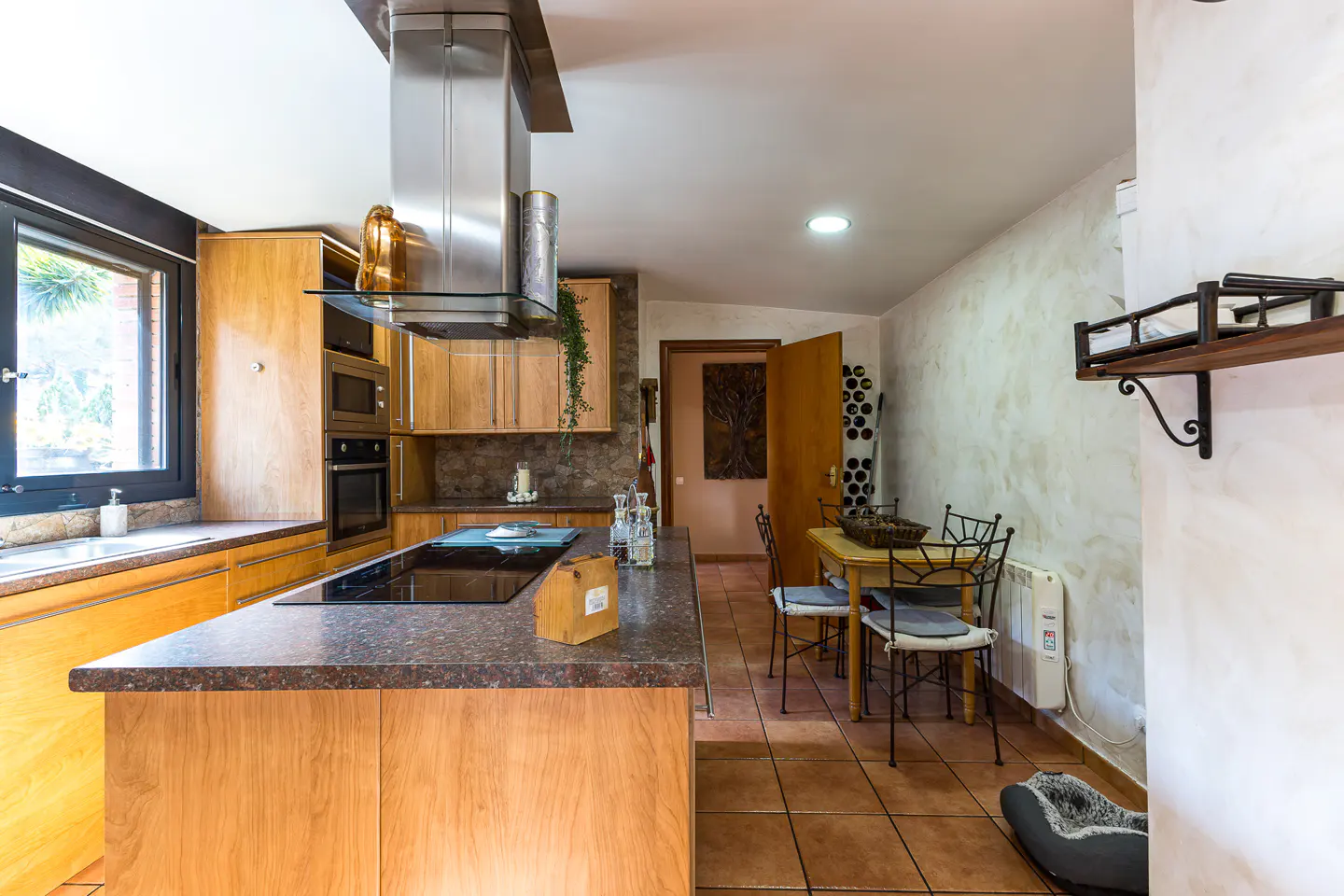 Modern kitchen with light wood cabinets, a dark granite island, stainless steel hood, and a small dining area with chairs.