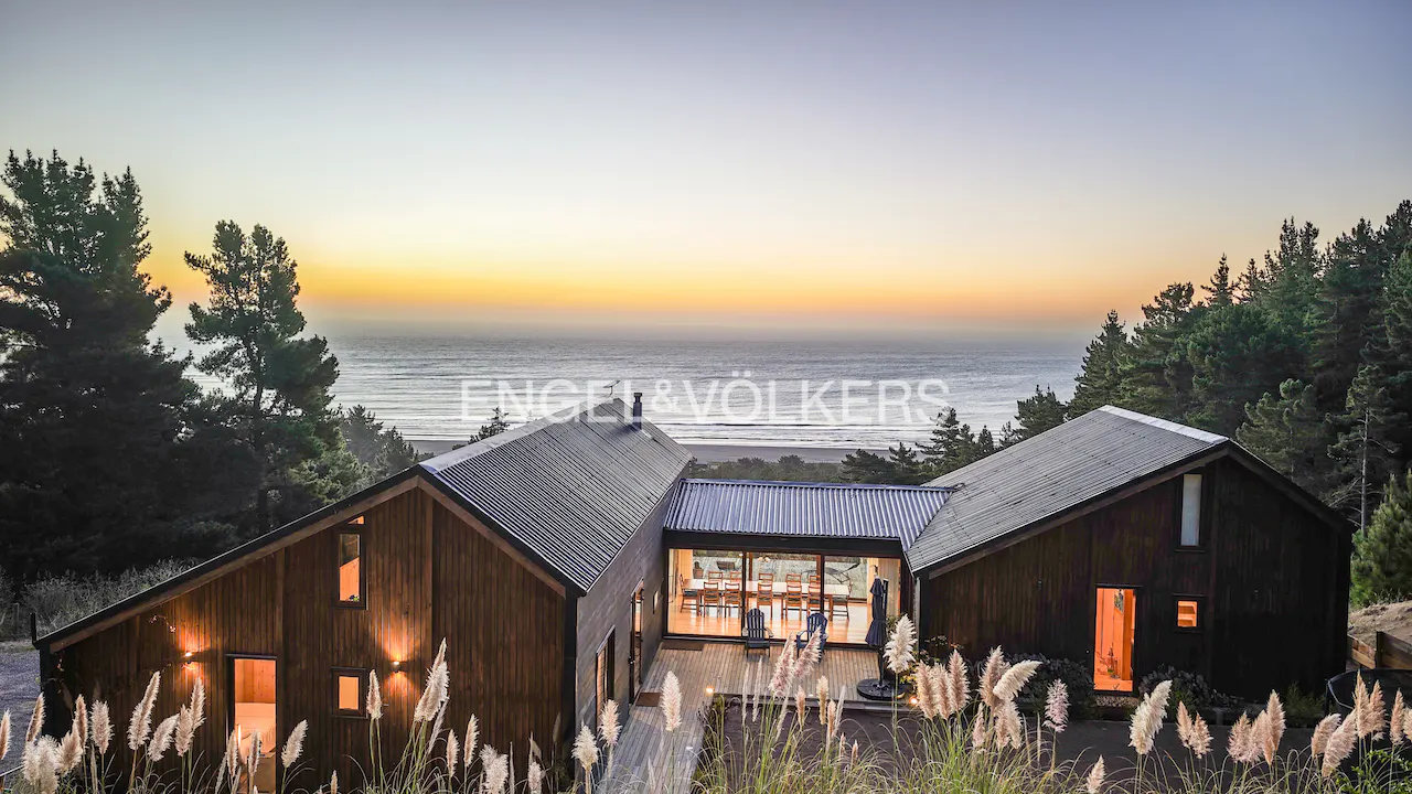 Exterior view of a modern, dark wood home with a metal roof, overlooking the ocean at sunset. Tall grass in the foreground.