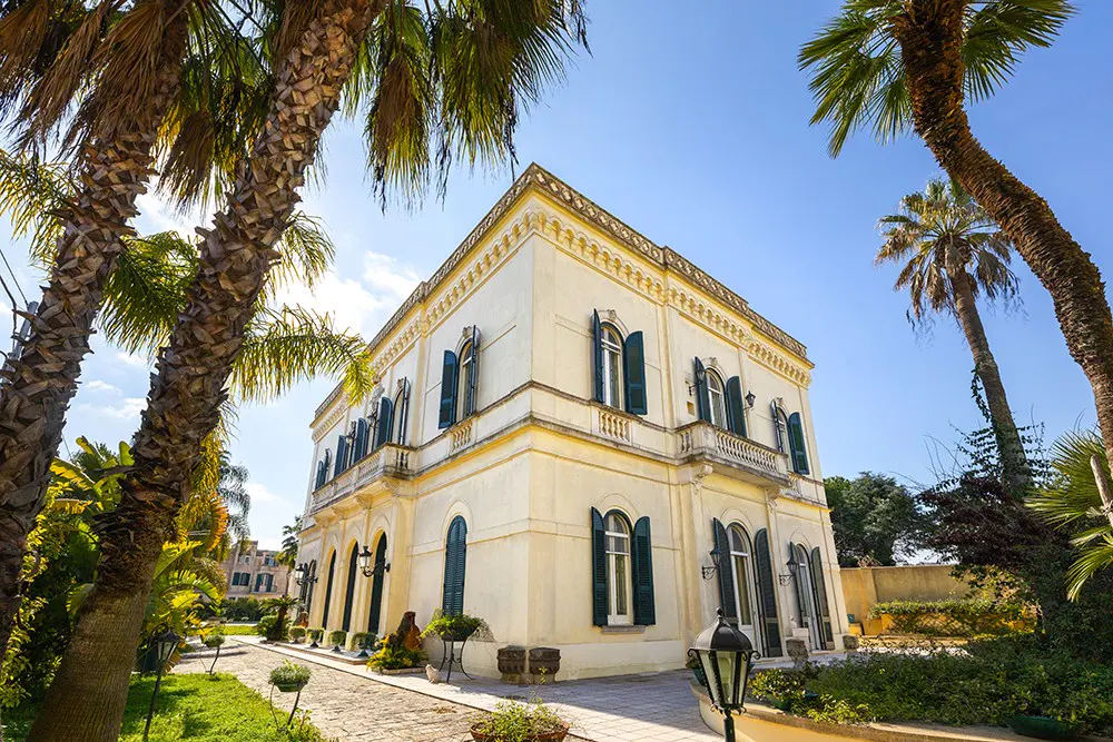 Elegant, cream-colored villa with green shutters, framed by palm trees against a clear blue sky.