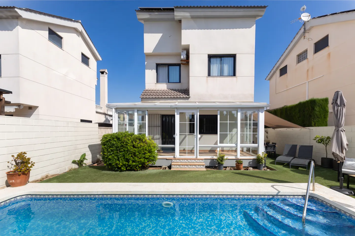 Backyard of a three-story white house with a swimming pool, glass-enclosed patio, green lawn, and lounge chairs.