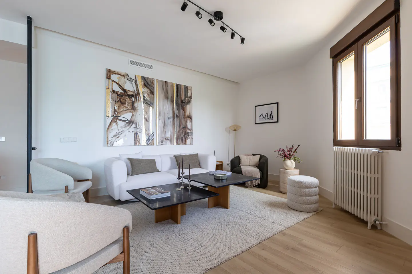Bright living room with white walls, wood floors, and a large abstract painting. Features a white sofa, armchairs, and a black coffee table on a light rug.