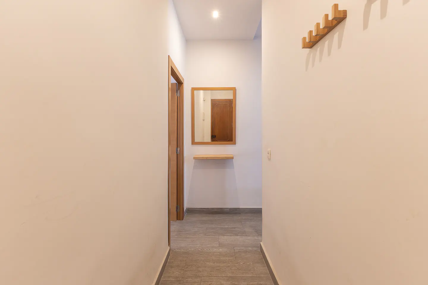 Bright hallway with light walls, wood-trimmed mirror, floating shelf, coat rack, and grey tiled floor.