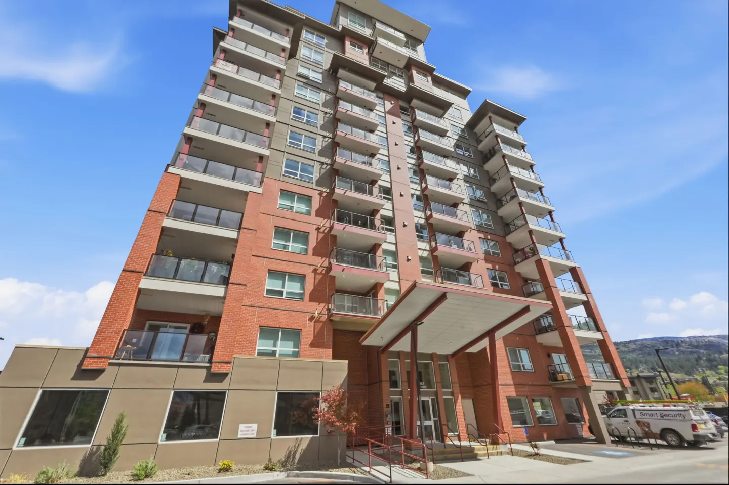 Exterior view of a tall, modern apartment building with balconies, red brick accents, and a covered entrance. Blue sky background.