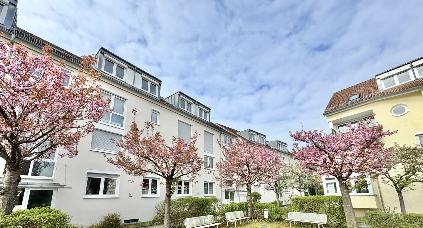 Exterior view of a light-colored apartment building with pink flowering trees, benches, and a cloudy sky.