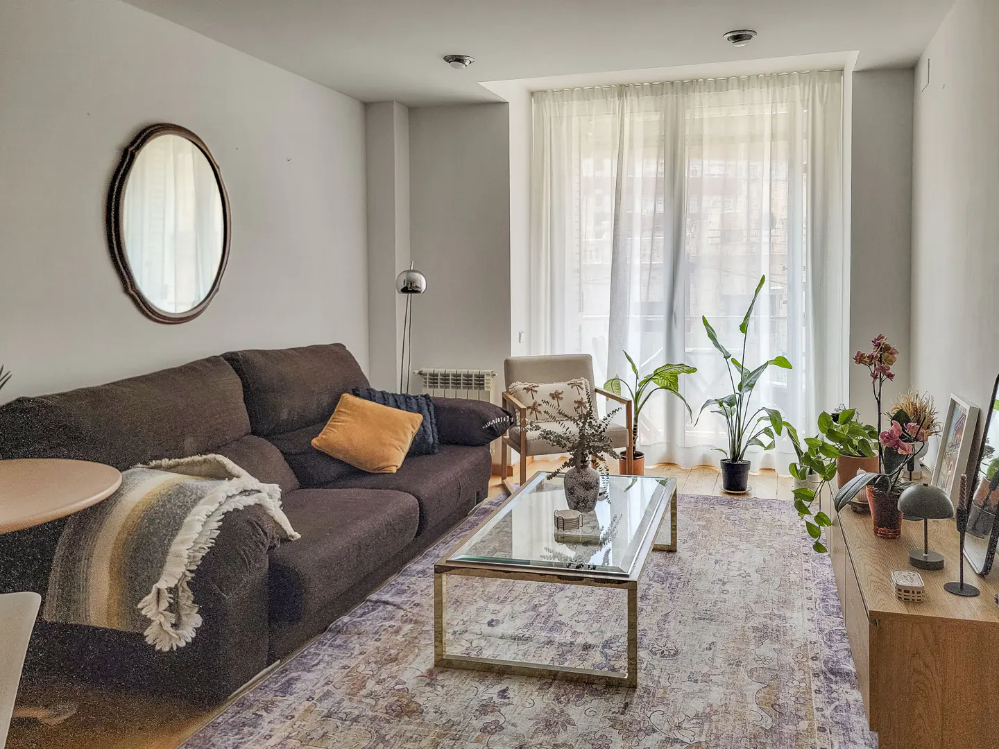 Living room with brown sofa, glass coffee table, and plants near sheer curtains. Oval mirror hangs on the wall.