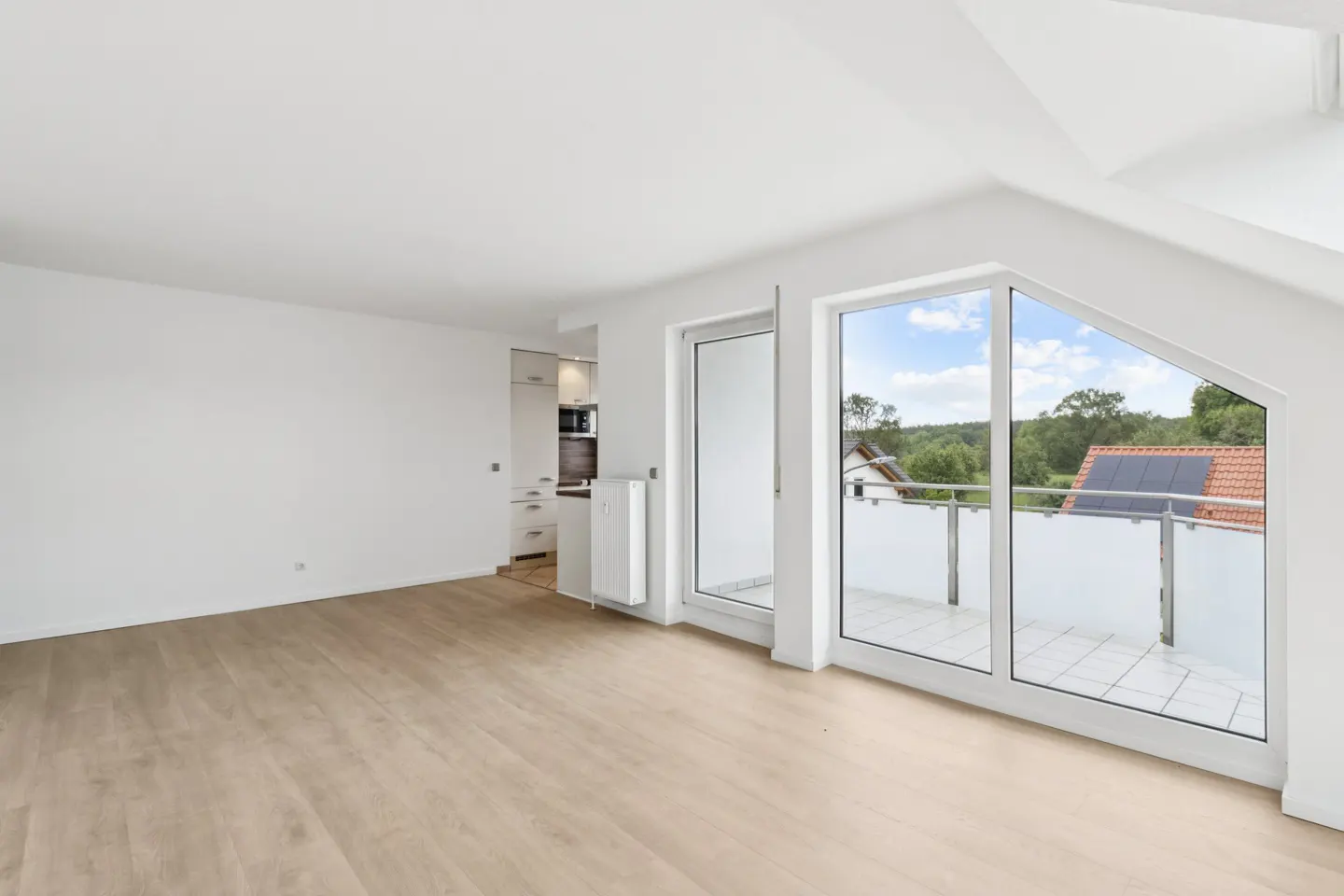 Bright, empty room with light wood floors, white walls, and a balcony with a view of trees and a house with solar panels.