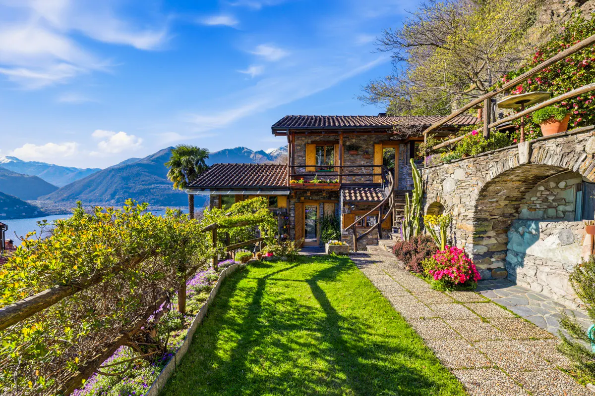 Stone house with a brown tile roof, green lawn, and stone path leading to the entrance. Mountains and a lake are in the background.