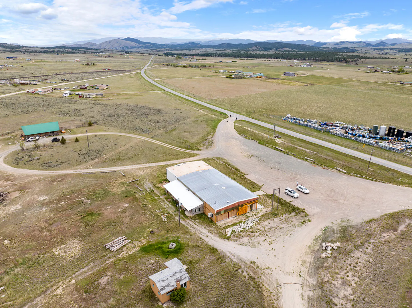 Aerial view of a rural property with a large commercial building, a small house, and distant mountains under a blue sky.