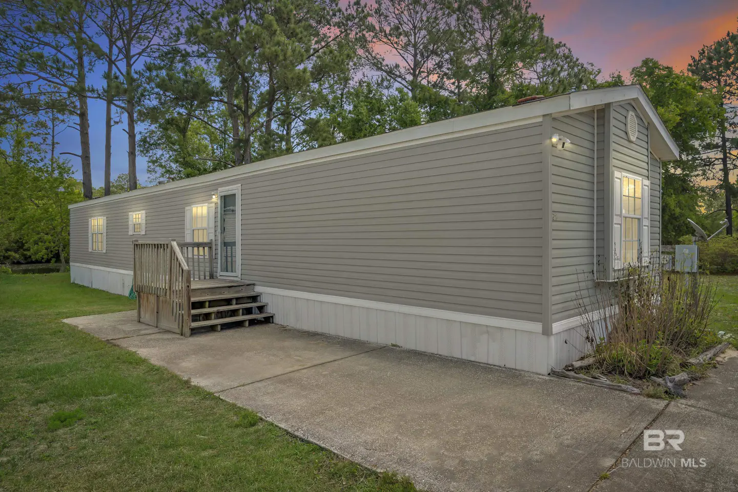 Exterior view of a gray mobile home with a wooden porch and steps, set against a backdrop of trees and a colorful sunset sky.