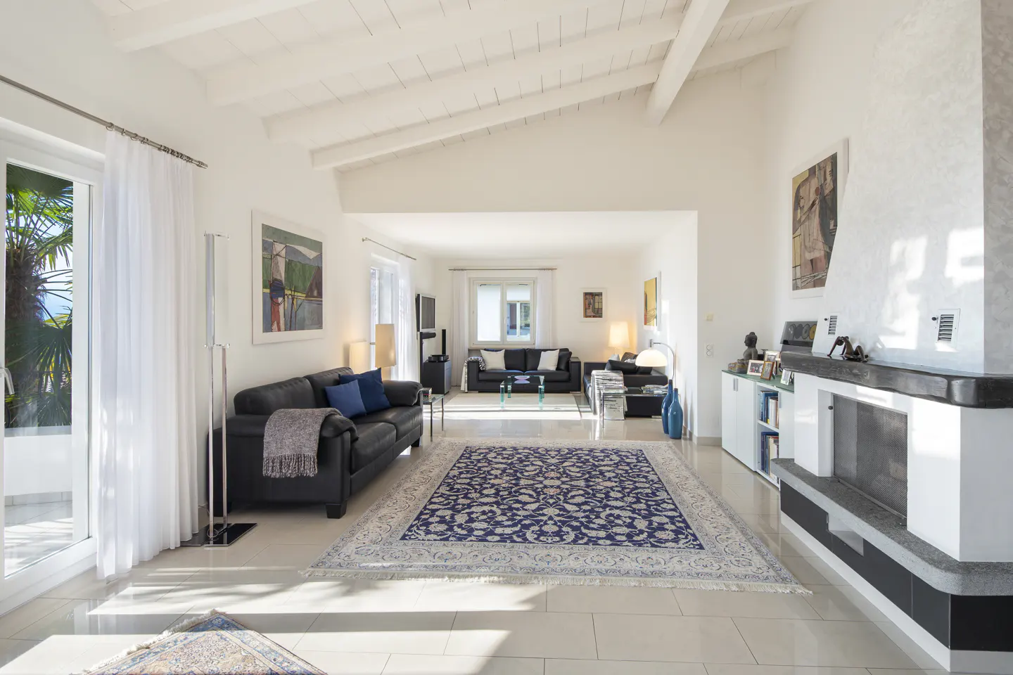 Bright, open living room with white walls, beamed ceiling, and tile floor. A blue patterned rug sits between two black leather sofas. A fireplace is on the right.