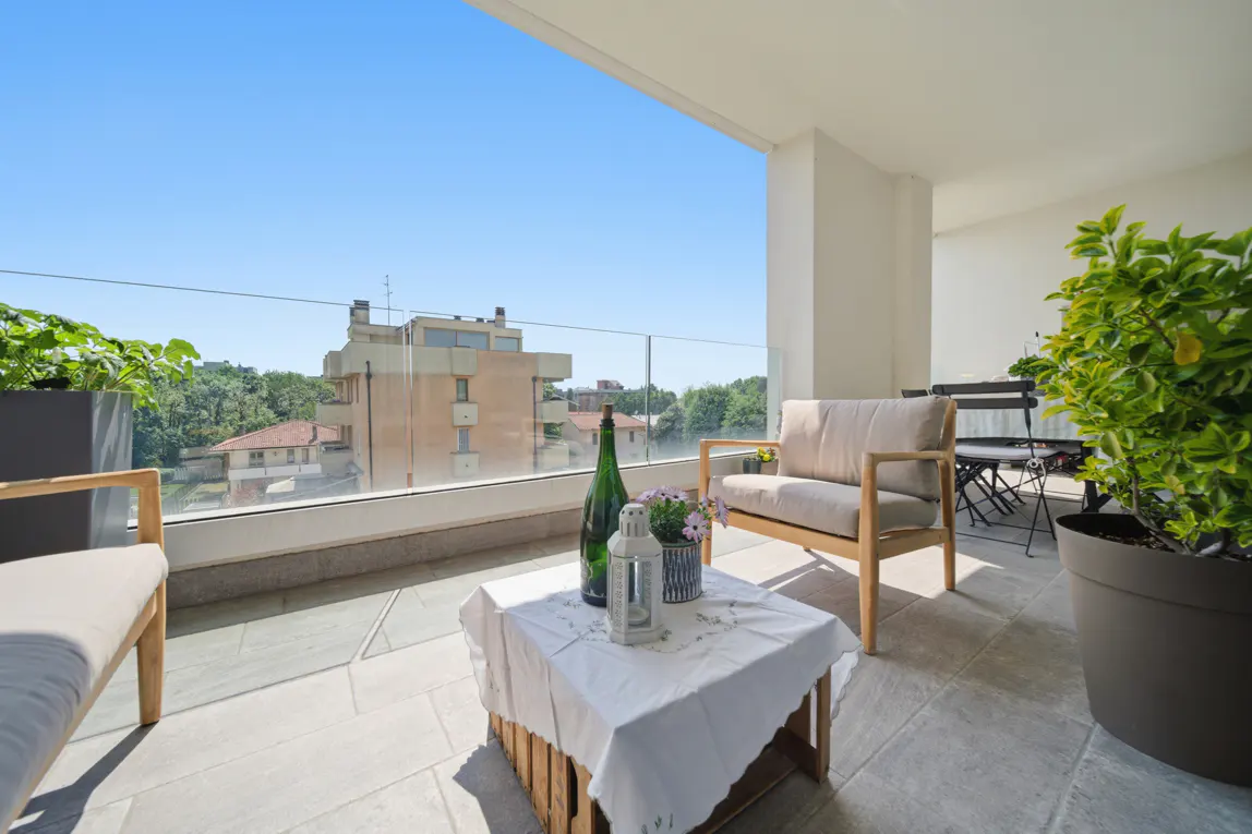 A bright balcony with stone floors, glass railings, and city views. A table with a white cloth, bottle, and flowers sits near a beige armchair.