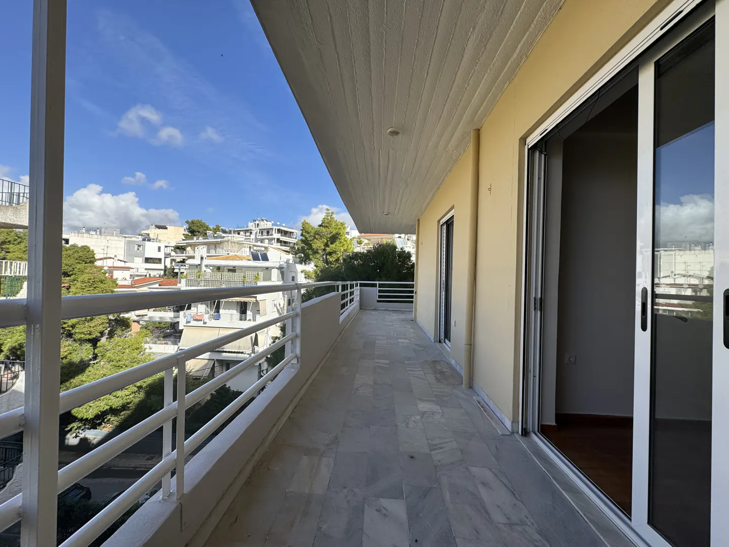 A long balcony with white railings overlooks a city with trees under a blue sky. Sliding glass doors lead inside.