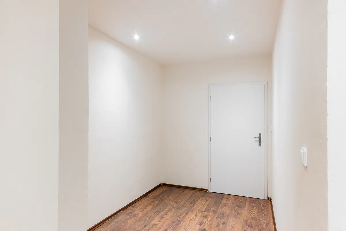 An empty room with white walls, a brown wood floor, and a white door. Two recessed lights are in the ceiling.