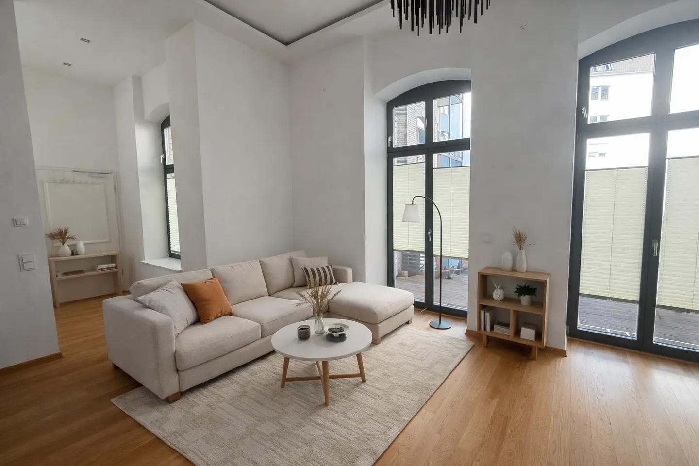 Bright living room with a beige sectional sofa, round coffee table, and large windows with blinds. A floor lamp and wooden shelf add to the decor.