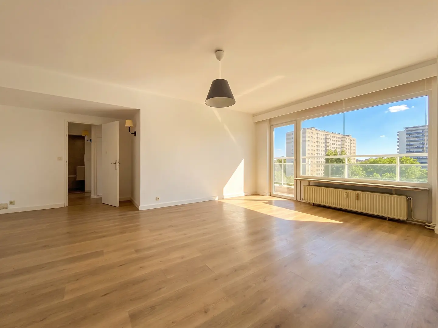 Bright, empty room with wood floors, white walls, and a large window showing city buildings and blue sky. A gray pendant light hangs from the ceiling.