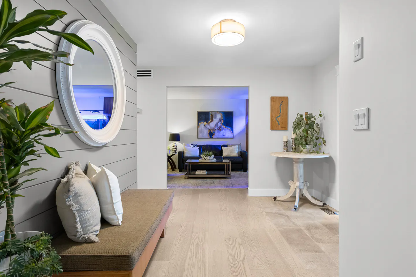 Entryway with a bench, round mirror on gray shiplap wall, and a view into a living room with a sofa and blue lighting.