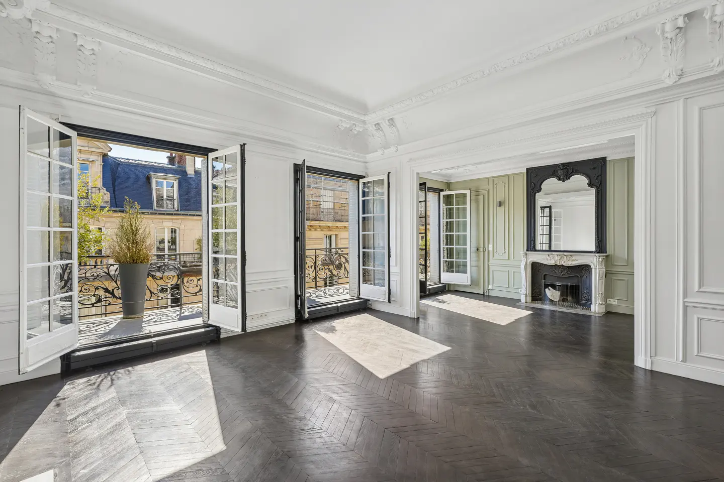 Bright, empty room with dark wood floors, white walls, and open French doors to balconies with city views. Fireplace with mirror in the background.