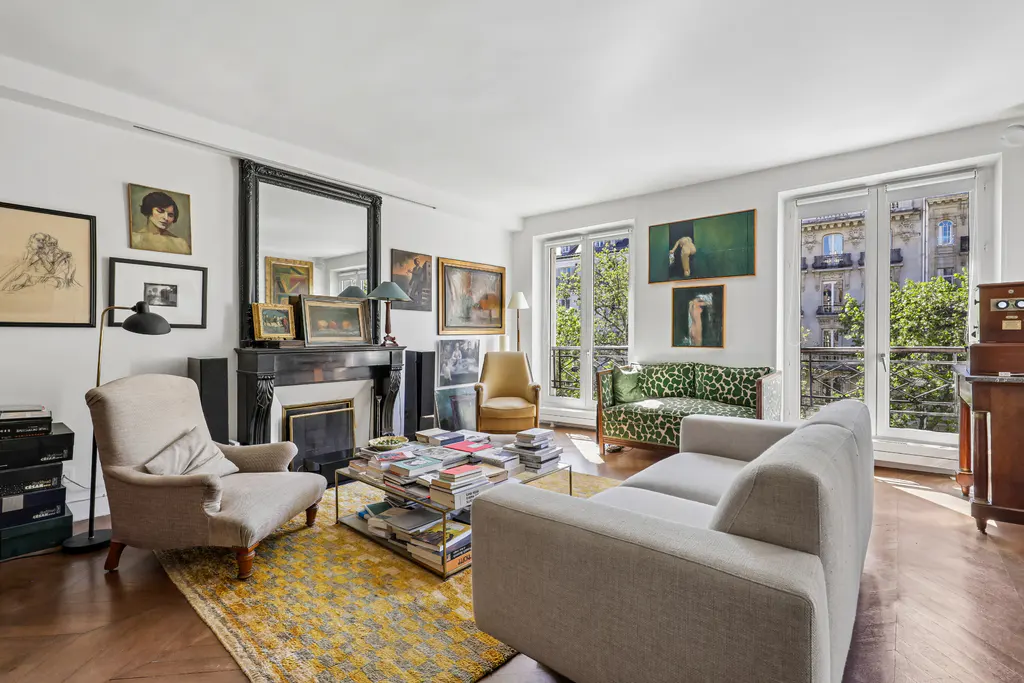 Bright living room with a fireplace, art, and large windows overlooking a city street. A sofa and chairs provide seating.