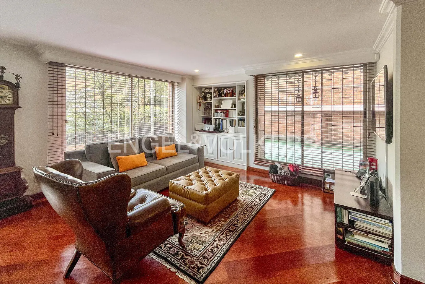 Living room with hardwood floors, gray sofa with orange pillows, leather chair, and ottoman on an ornate rug. Windows with blinds and a built-in bookcase.