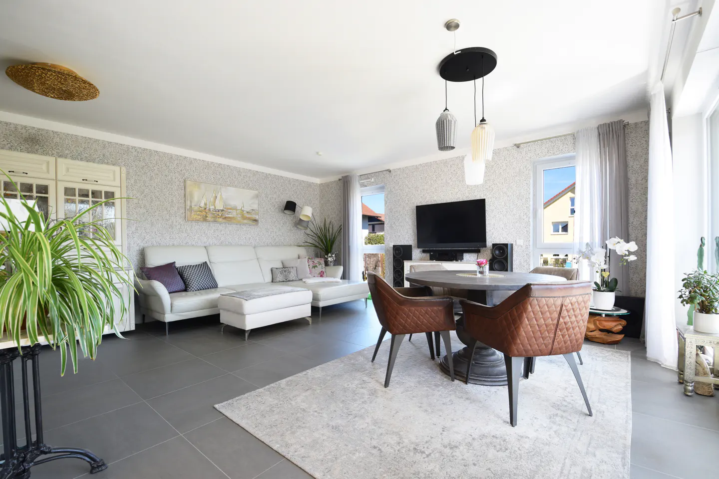 Bright living room with white sofa, brown leather chairs around a round table, and a large TV. Gray tile floor and patterned wallpaper.