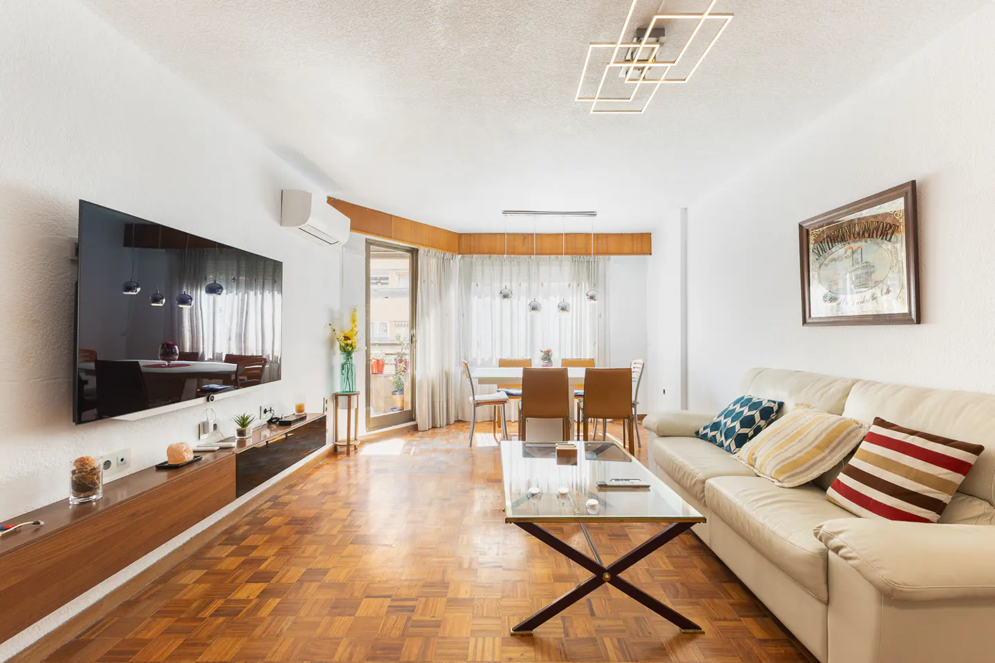 Bright living room with parquet floors, white walls, and a cream sofa with colorful pillows. A glass coffee table sits in front of the sofa. A large TV is mounted on the wall.