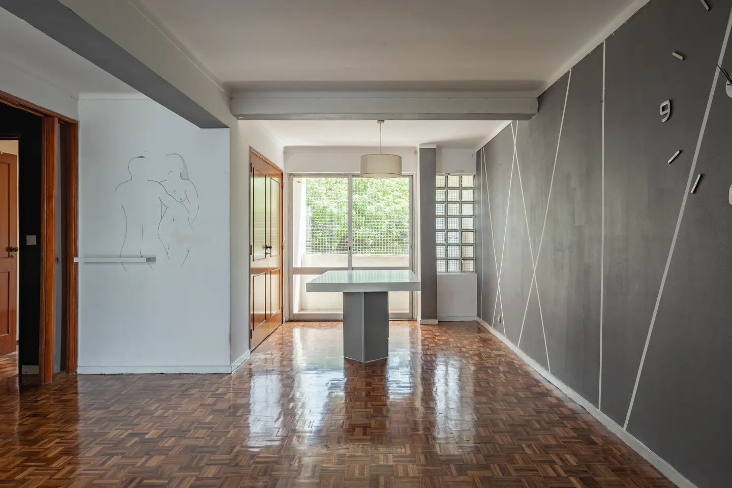 An interior shot of a room with parquet floors, a gray wall with white stripes, and a white wall with a drawing.
