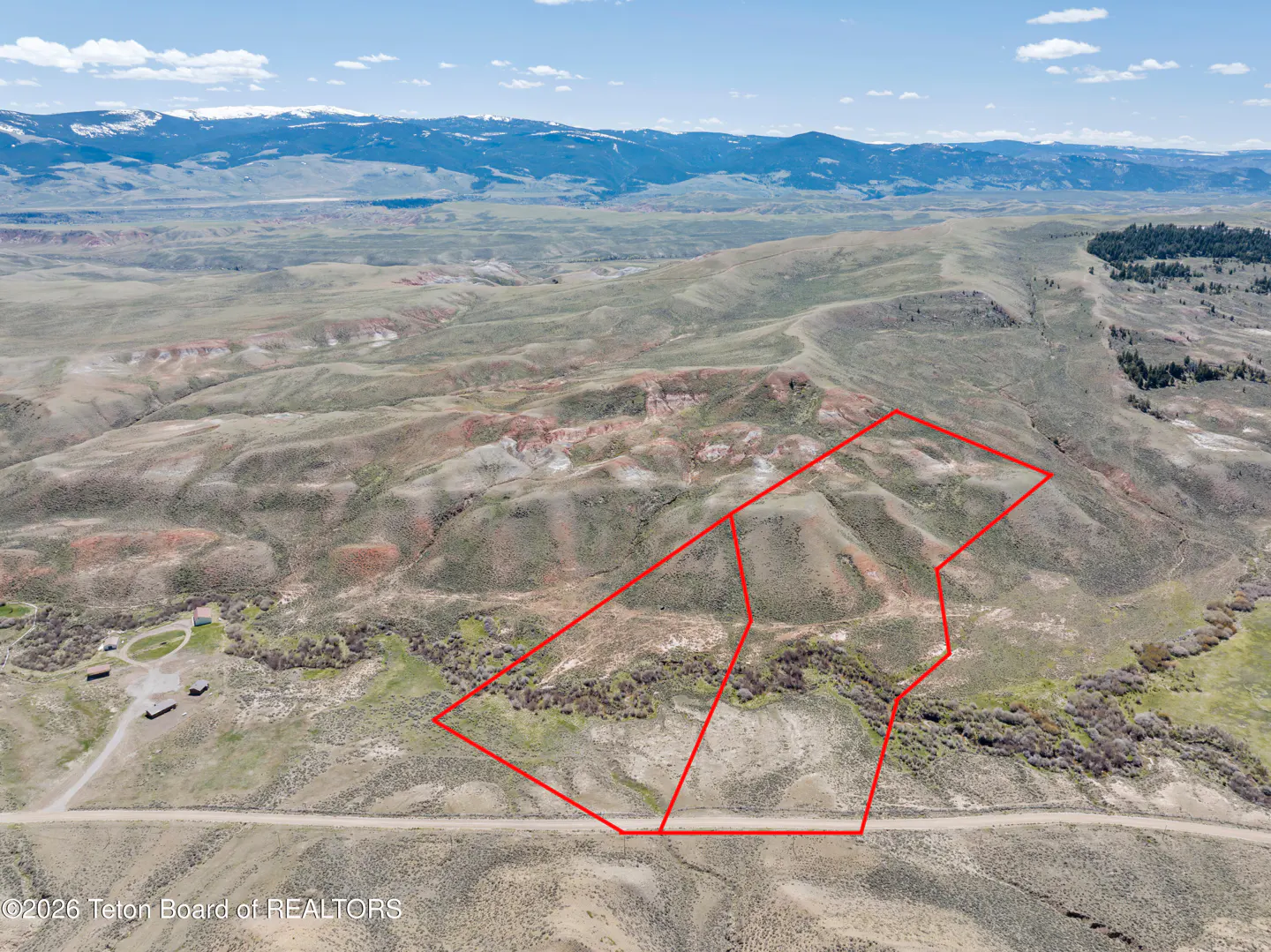 Aerial view of a large, hilly plot of land outlined in red, set against a backdrop of distant snow-capped mountains.