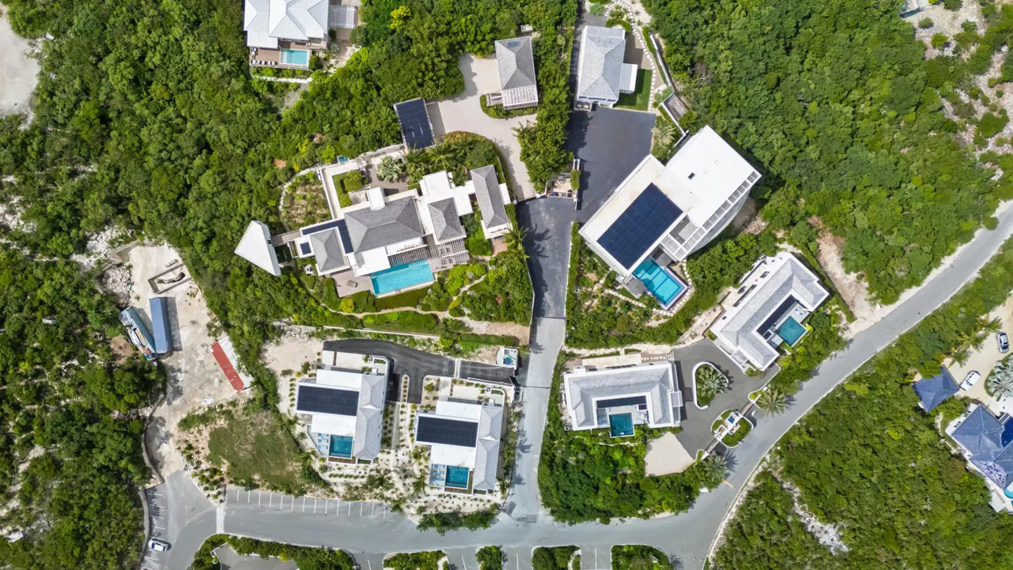 Aerial view of luxury homes with pools and solar panels nestled in lush green vegetation. Roads and parking areas are visible.