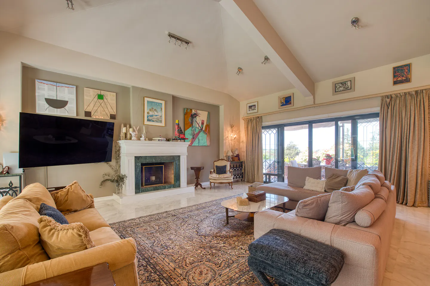 A spacious living room with tan sofas, a marble fireplace, and a large patterned rug. Sliding glass doors lead to an outdoor area.