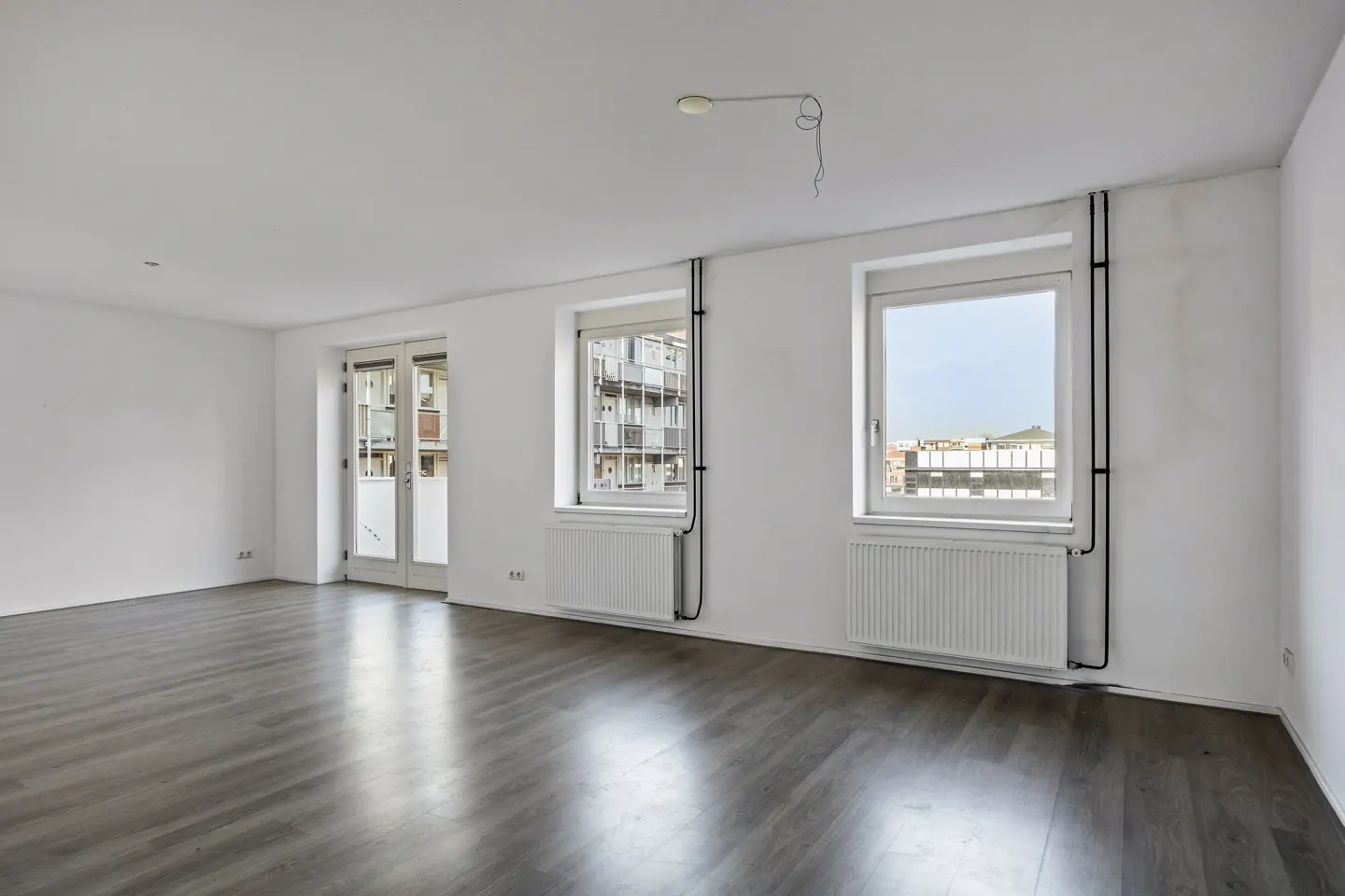 Bright, empty room with gray wood floors, white walls, two windows, and a glass door. Radiators are under the windows.