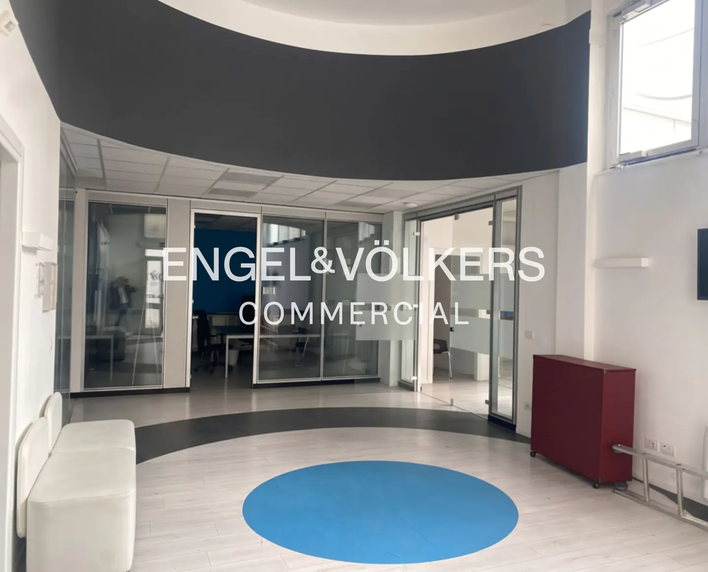 Bright office space with glass-walled offices, a blue circle on the floor, and a white bench. "Engel & Völkers Commercial" logo.