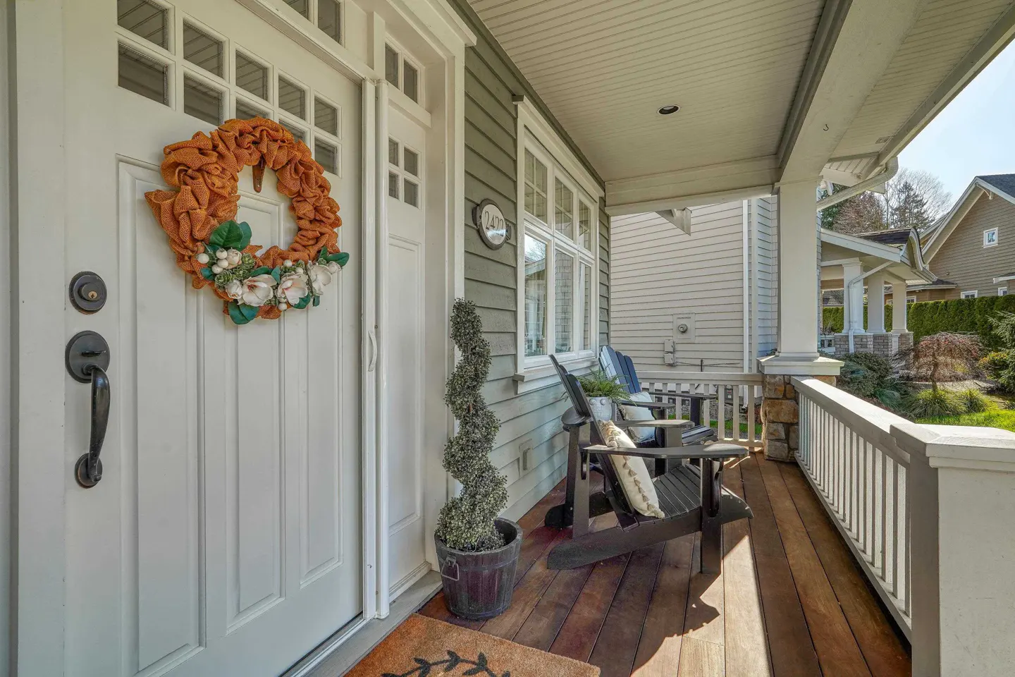 Front porch of a house with a white door, orange wreath, and wooden chairs. The house has green siding and white trim.
