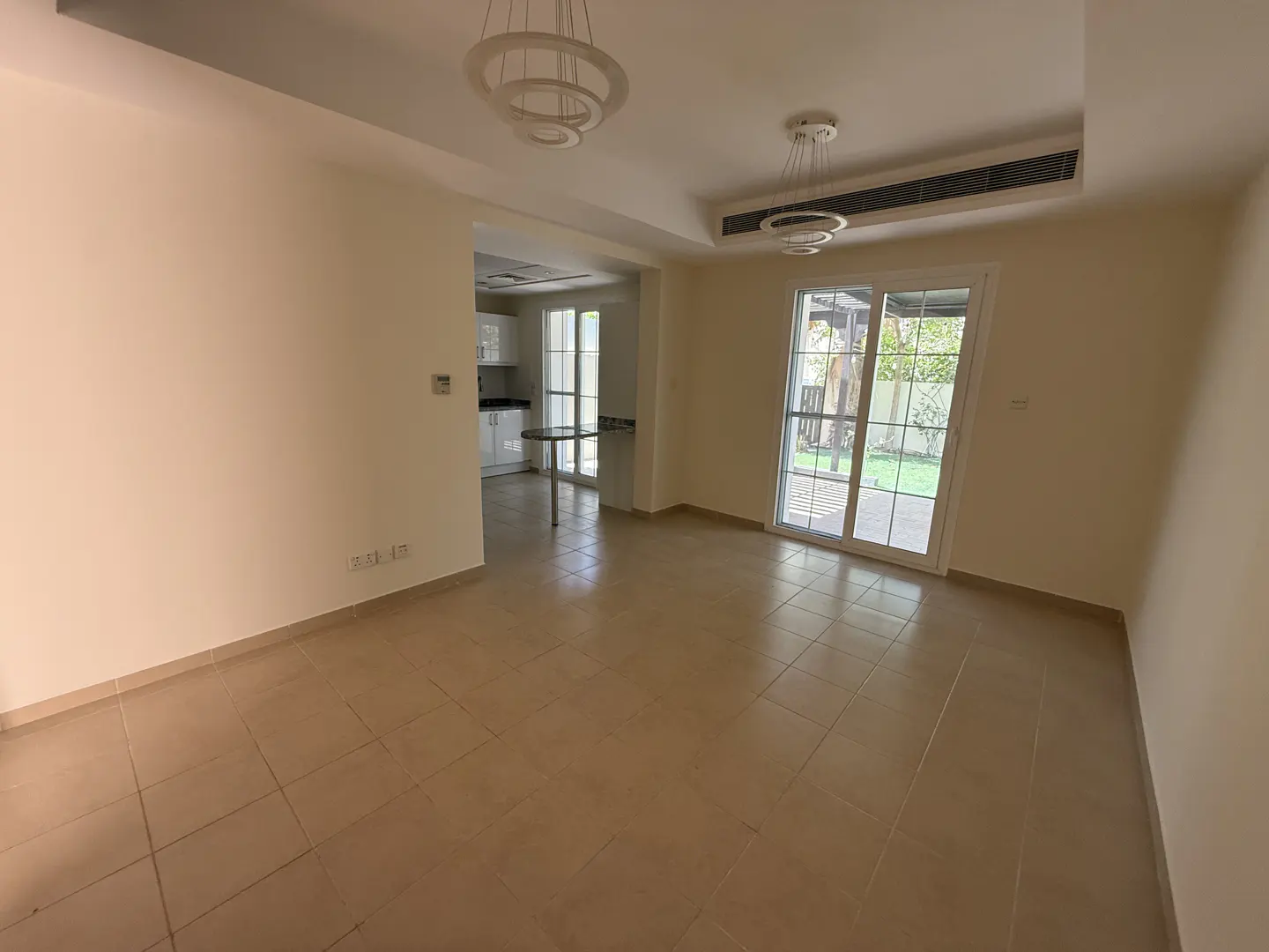 A bright, empty living room with beige tile floors, white walls, and sliding glass doors to a green yard. Two modern ring chandeliers hang from the ceiling. A kitchen is visible in the background.