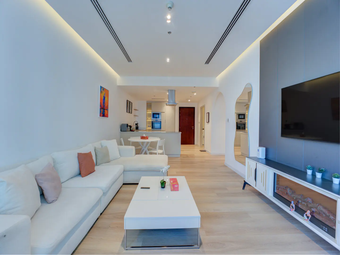 Bright, modern apartment interior with white sectional sofa, coffee table, TV, and open kitchen in the background.