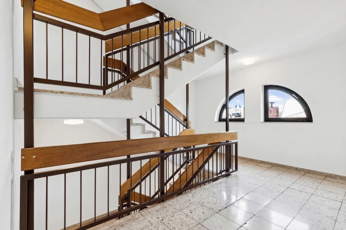 Interior view of a stairwell with brown railings, stone stairs, and white walls. Two arched windows are visible on the right. The floor is tiled.