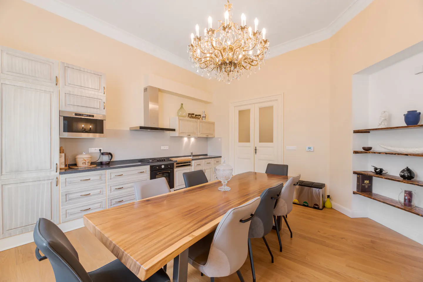 A bright kitchen and dining area with a wood table, gray chairs, and a crystal chandelier.