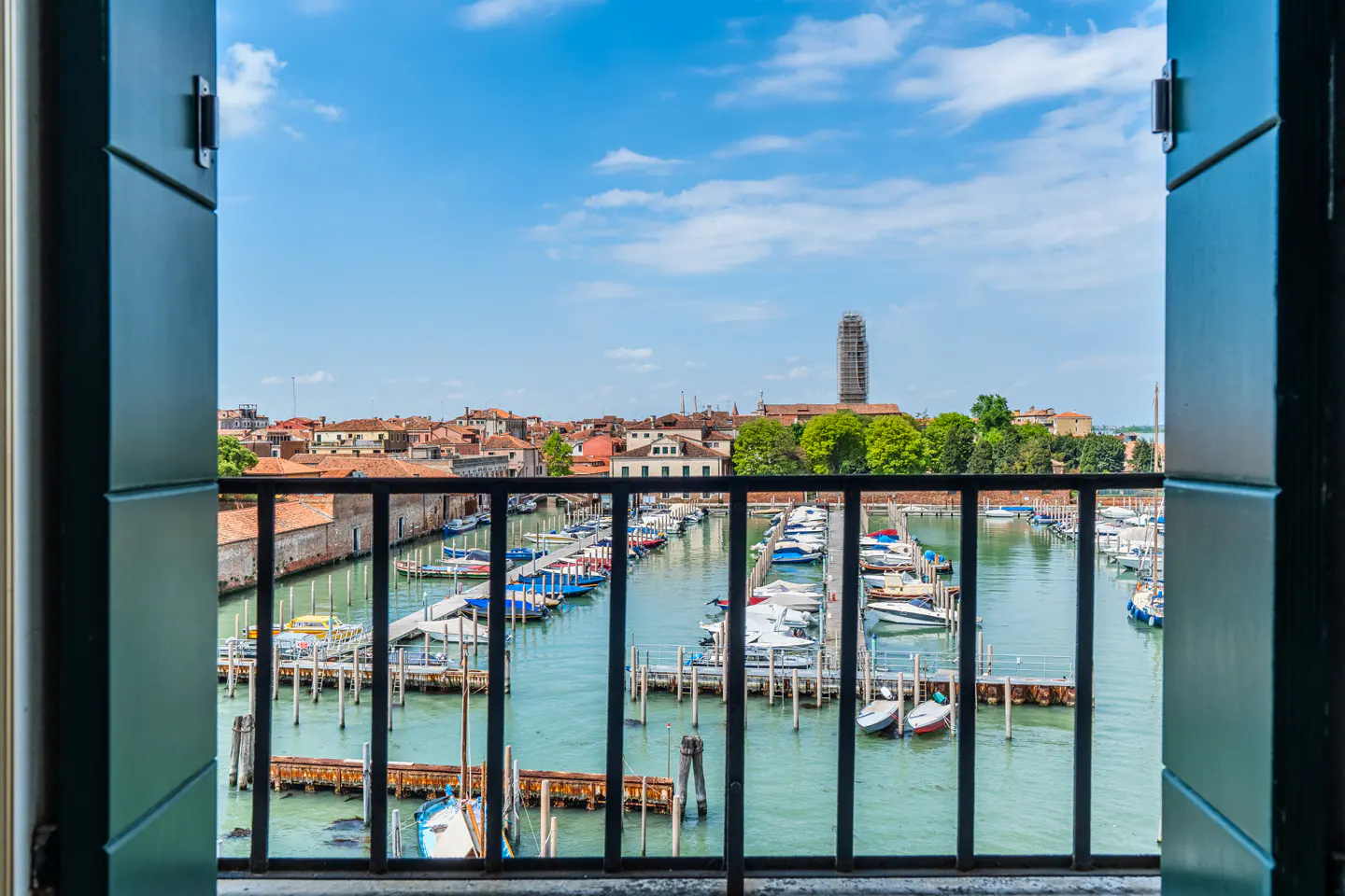 View from open window with green shutters of Venice marina filled with boats, buildings, and blue sky.