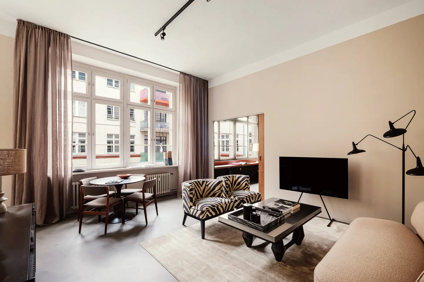 Modern living room with zebra-print armchairs, a black coffee table, a TV, and a dining area by large windows with curtains.