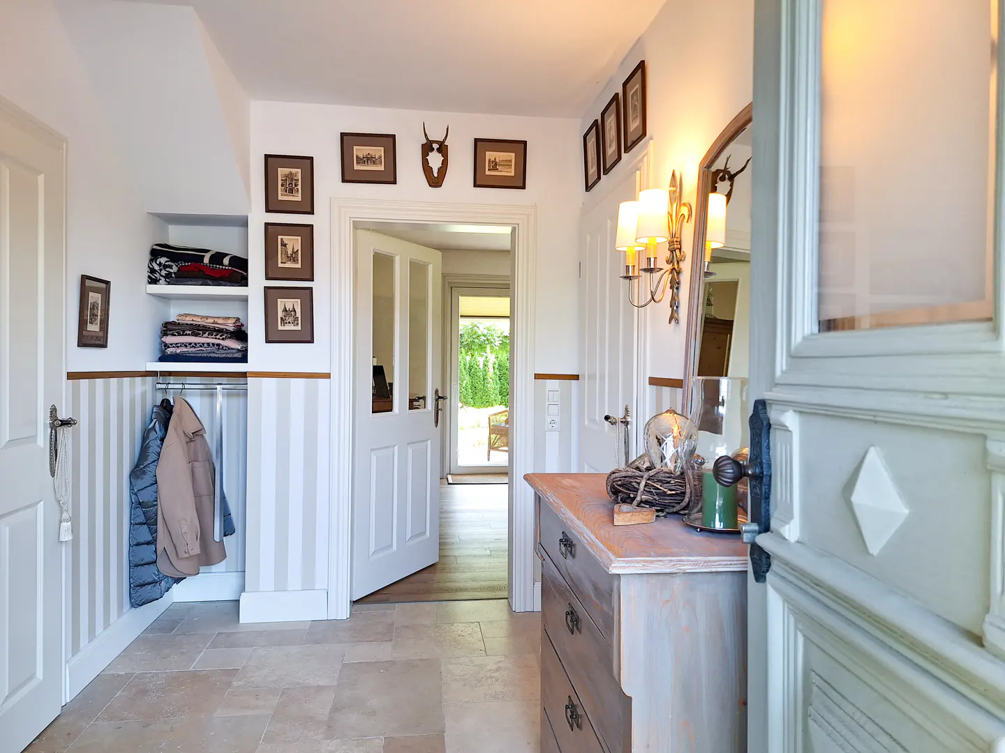 Entryway with stone floors, striped walls, and white trim. A coat rack and shelves hold jackets and blankets. Framed pictures and antlers decorate the walls.