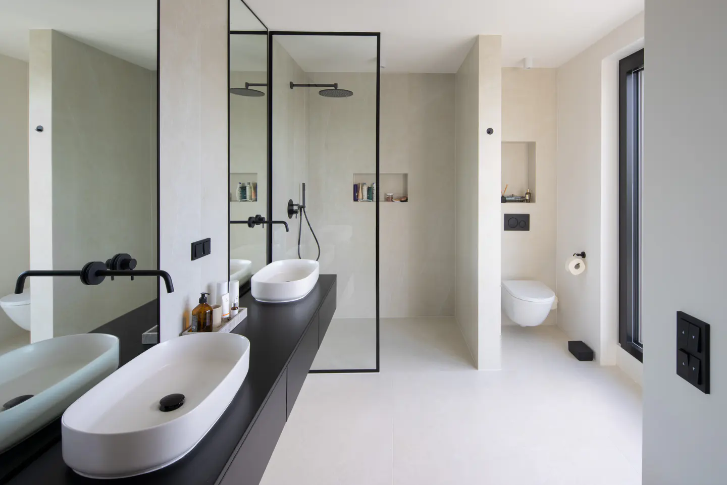 Modern bathroom with white walls, black fixtures, and white oval sinks on a black vanity. A glass-enclosed shower and toilet are visible.