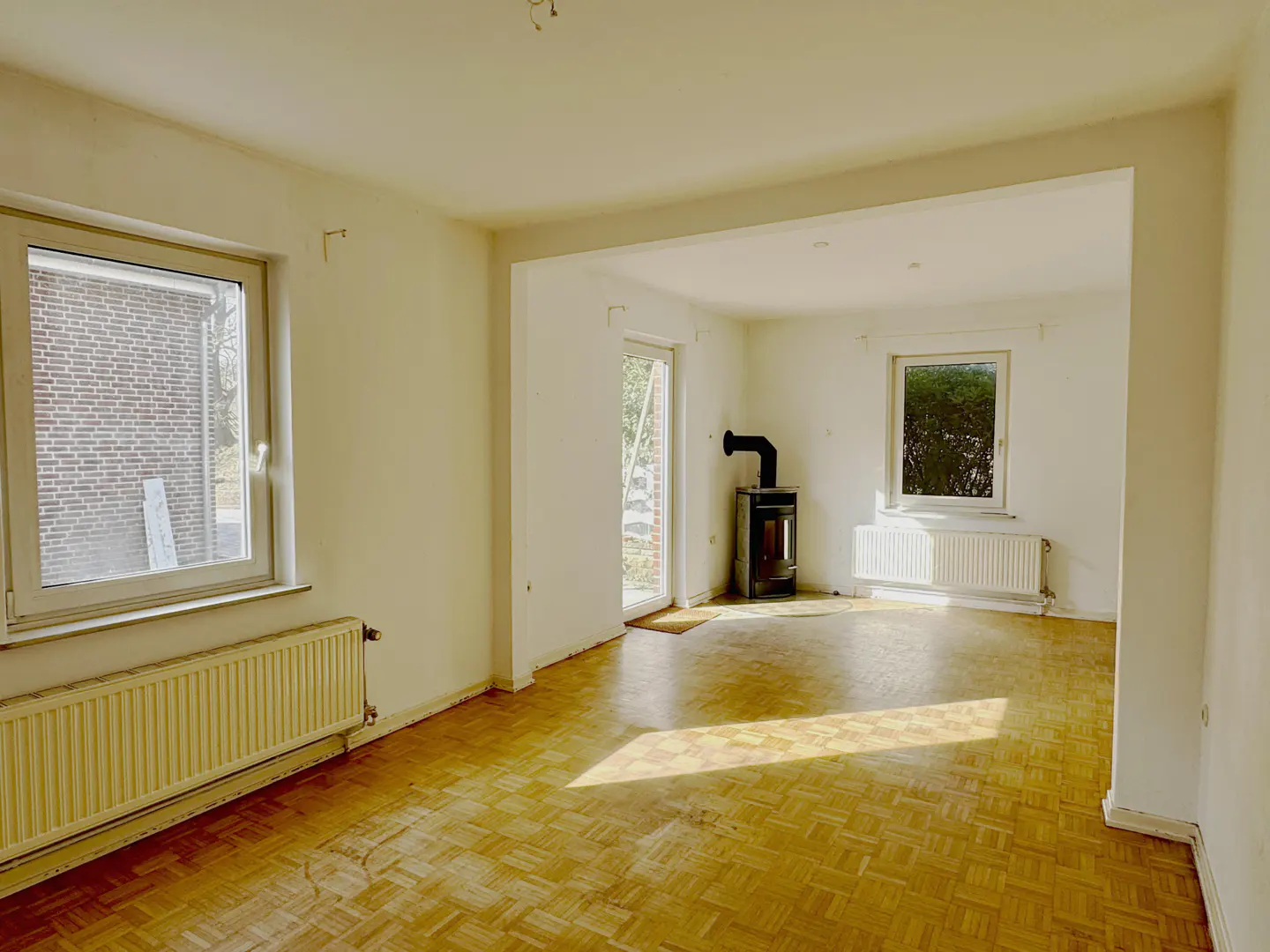Bright, empty living room with parquet floors, white walls, and two windows. A black wood-burning stove sits near a glass door. Radiators under windows.
