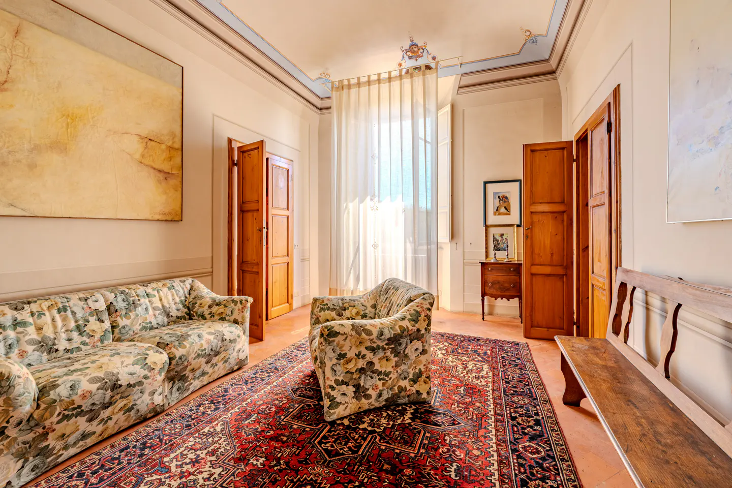A sunlit room with floral sofa and armchair, a large red patterned rug, wooden doors, and a wooden bench on terracotta floors.