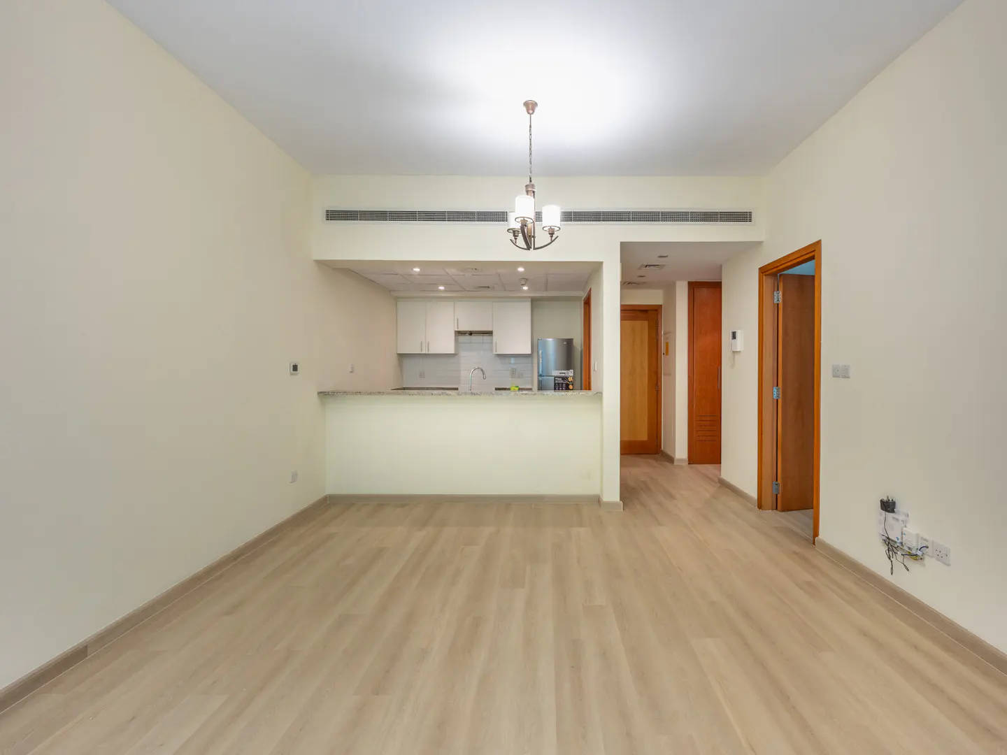 An open-concept living space with light wood floors, cream walls, and a kitchen with white cabinets and stainless steel appliances. A chandelier hangs from the ceiling.