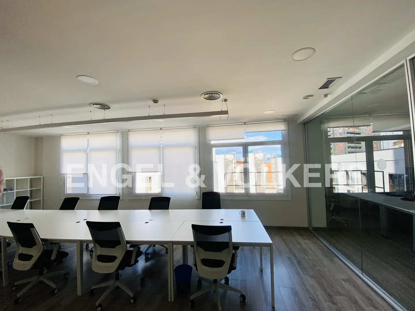 Bright office space with a long white table, black chairs, and large windows with roller shades. A glass-walled office is on the right.