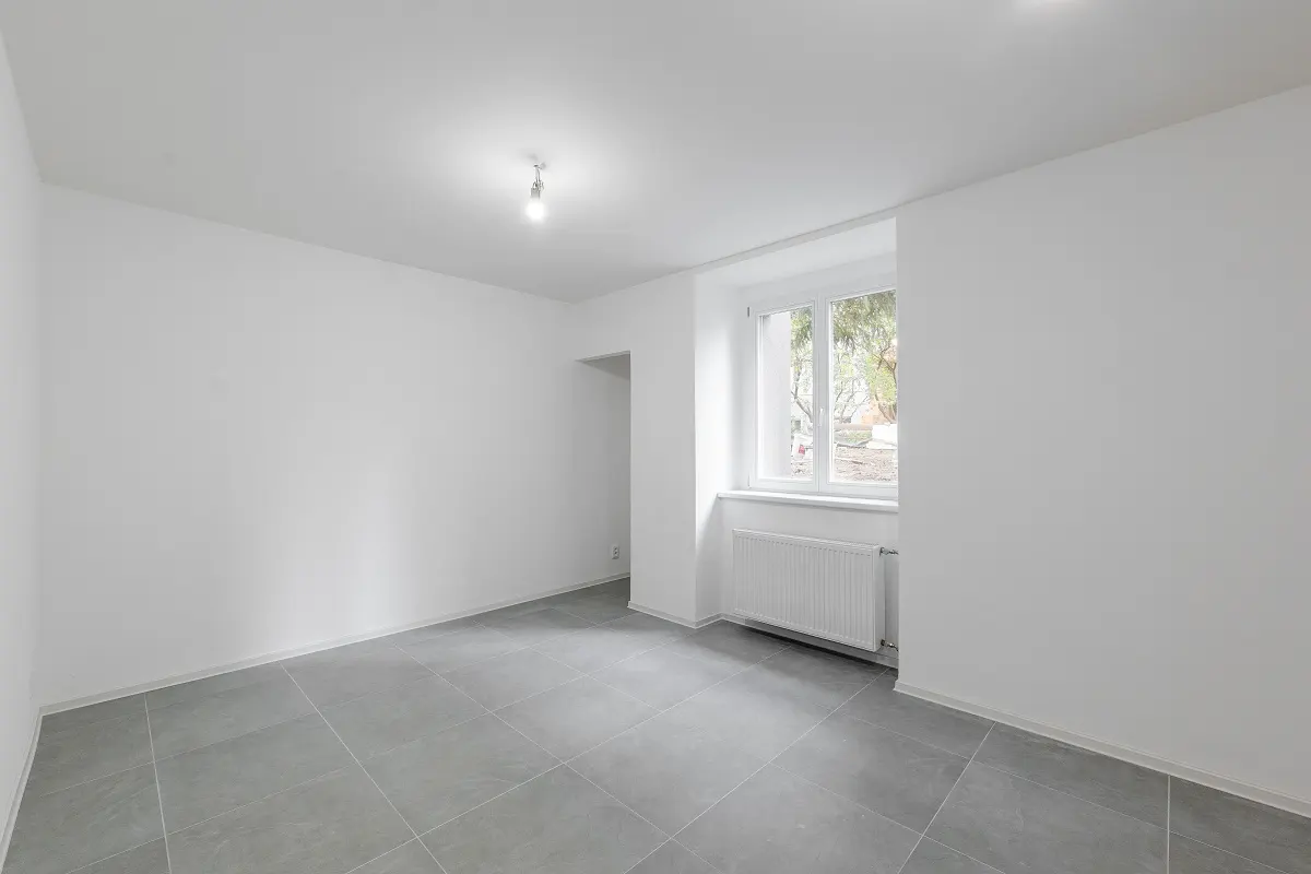 An empty, bright room with white walls, gray tile floor, and a window with a white frame. A white radiator is under the window.