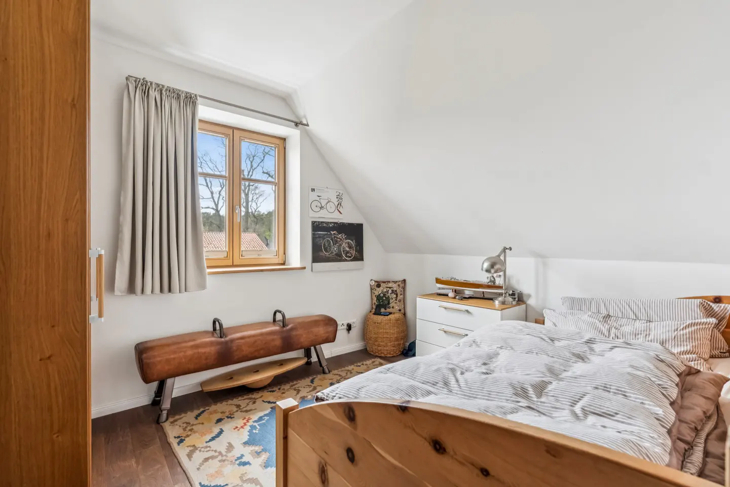 Attic bedroom with a wooden bed, white walls, and a window with gray curtains. A pommel horse sits near the window. A colorful rug is on the floor.