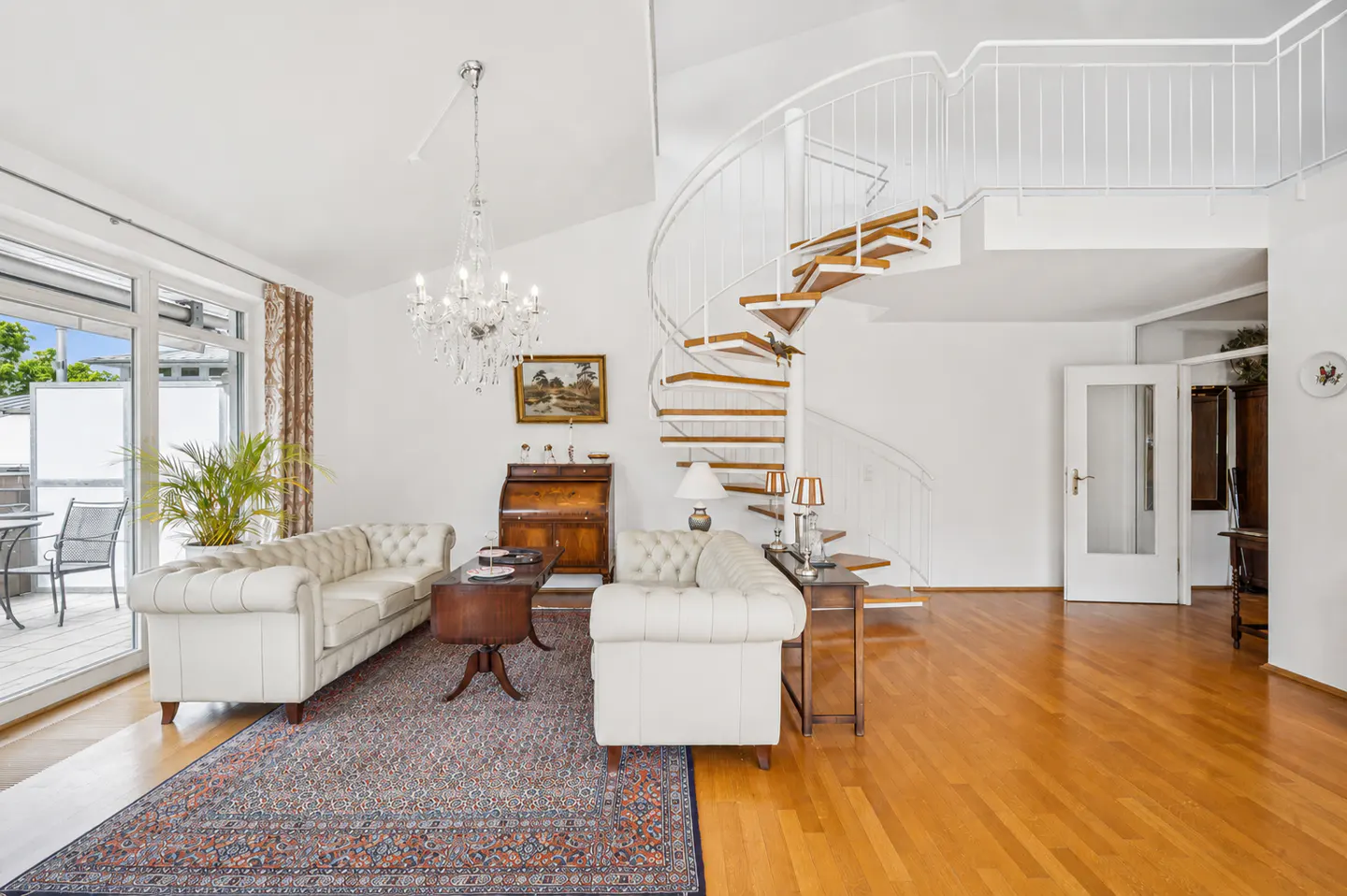 Bright living room with white tufted sofas, a patterned rug, a crystal chandelier, and a white spiral staircase.