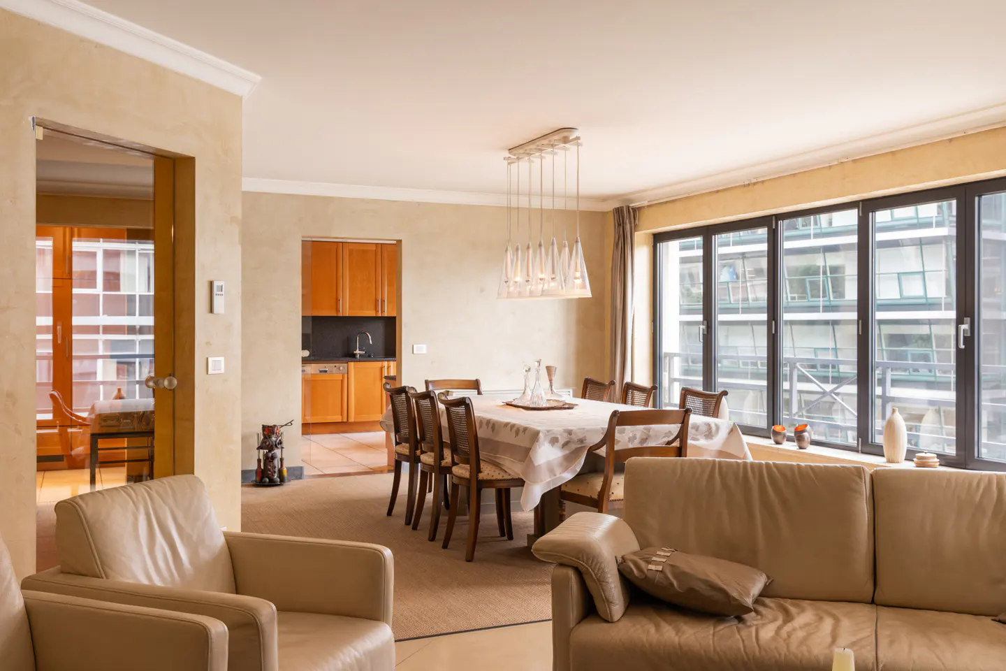 Living room with beige leather sofa and chairs, dining table with chandelier, and kitchen view. Large windows offer city views.
