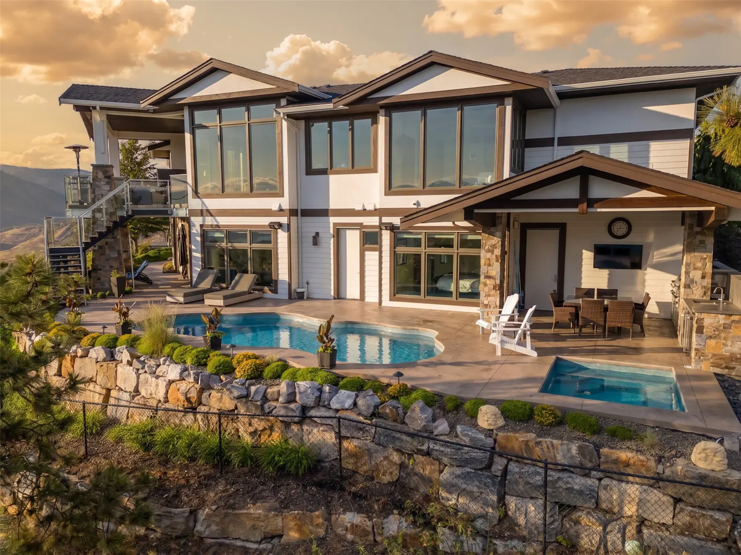 A modern two-story white house with a pool, hot tub, and outdoor kitchen on a stone-terraced lot. Large windows reflect the sky.