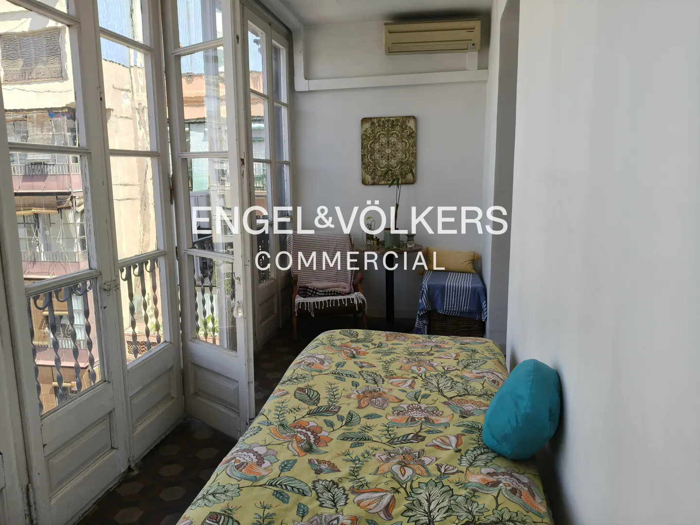 Bedroom with yellow floral bedspread, blue pillow, chair, and open balcony doors. Engel & Völkers logo overlay.